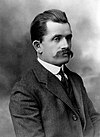 Black and white photo of a skinny young man with a large moustache, frowning with an neutral expression on his face. He is wearing a dark grey suit paired with a white undershirt, and a tie.