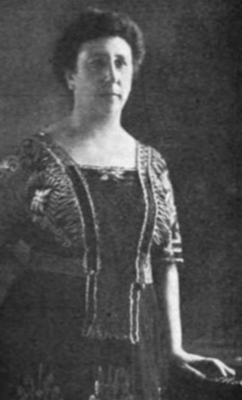 A white woman, standing, wearing a dark dress with a square neckline