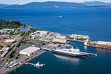 One large ship and one small ship are docked at a port. There is a large expanse of water with another small ship approaching and mountains in the background.