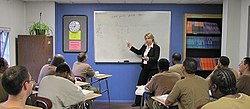 Une femme se tient devant une salle de classe et indique des fractions sur un tableau blanc. Onze hommes sont assis à des bureaux en face d'elle.