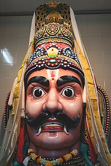 A big moustached male head, with big eyes, big ears and thick eyebrows. Fangs protrude from the sides of his mouth. The head wears a conical crown, with a cobra hood at the top. A floral garland and gold necklace are seen around the neck.