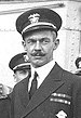 Head and shoulders of a white man with a handlebar mustache, wearing a peaked cap and a dark jacket with two rows of ribbon bars on the left breast. Several other men in similar clothing can be seen behind him.