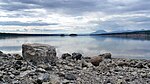 A wide flat river flows along a rocky shore toward a mountain range in the far distance.