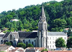 Photo représentant sur fond de verdure l'église Saint-Georges. Au centre, son clocher flèche et sur la gauche sa nef de laquelle ressort son transept proéminent. À droite du clocher, un long et haut bâtiment de trois étages situé derrière l'église donne l'impression d'une autre nef, presque symétrique.