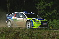 Voiture bleu foncé, verte et recouverte de terre sur les flancs, vue de trois-quarts.