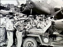 Approximately 15 men wearing military uniforms in discussion around a jeep, parked in front of a twin-engined aircraft