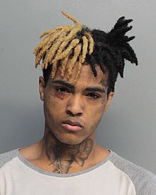 A sullen-looking young man against a white wall with an elephant tattooed on his neck and the right side of his hair bleached