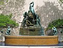 An outdoor fountain with water passing from one plate two another until it ends up in the fountain base. The fountain has several women holding plates and is backed by a structural wall. Trees are adjacent to the fountain on either side.