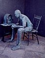 Un uomo siede su una sedia, senza poggiare la schiena sullo schienale e con i gomiti poggiati sulle ginocchia. La scultura è The Fireside Chat di George Segal (Franklin Delano Roosevelt Memorial, Washington).