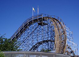 The first drop and returning track is observed from the viewpoint on the ground. The drop is prominently featured with the returning track in the background. The empty track curves to the left amidst foliage and support structures.