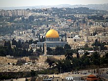 A city scene. Many of the buildings look ancient. In the centre, there is a large building topped by a golden dome. In the background, there are modern-looking high-rise buildings.