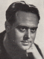 Publicity shot of a strong-featured, clean-shaven man in his 40s, leaning forward a little, with his chin on his hand.