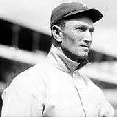 A man wearing a light colored baseball jersey and dark colored baseball cap looks into the distance.