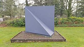 From the angle of this photo, the gray-painted metal sculpture appears to be a large rectangle, approximately 8 feet wide by 6 feet high. A triangular section juts out from the lower half (diagonally from top left to bottom right, 0 inches to 2 feet, respectively) kind of like the corner of a pyramid.