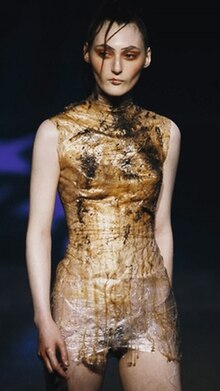 A woman wearing a short sleeveless dress made of plastic, smeared with filth