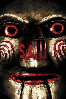 The box art features the face of an antique-looking Billy doll. The face is gold in color and is shiny. It has red eyes and red lips. The word "Saw" in red letters is in the center.