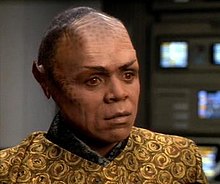 The dark-skinned Tom Wright is in makeup and costume as Tuvix. Black makeup is mottled across his cheeks and high brow, black straight hair is combed back from the hairline, pointed ear prosthetics have been applied, and amber-colored contact lenses are visible. The costume is colored in mustard and black like Tuvok's, but patterned in flowers and cut to a style like Neelix's customary clothing.