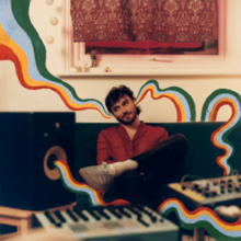 A man sitting in front of several keyboards and next to a speaker that has drawn waves of sound coming out of it that go through one of his ears and out the other.