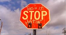 In the photo, a red stop sign can be seen. On the stop sing, the word "Stop" is in the center, and written above it are the words "This Is It" and at the bottom of the sign, the word "Hatin'" is written in capital letters; all the words on the sign are written in yellow. In the background, a blue sky, that is mainly full of white clouds, is shown and a metal poll is shown behind the sign.