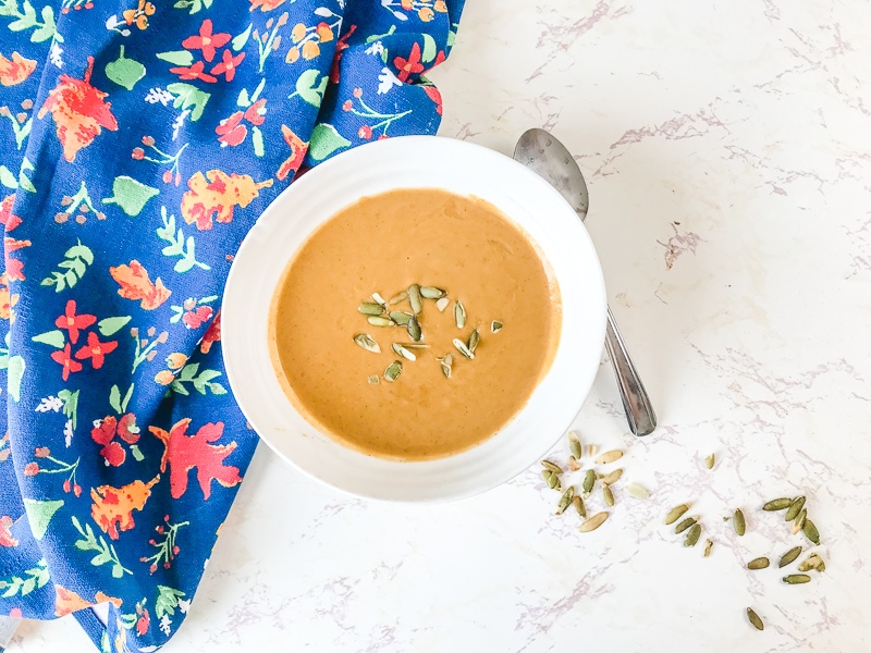 A white bowl filled with pumpkin soup next to a handful of pepitas on the counter.