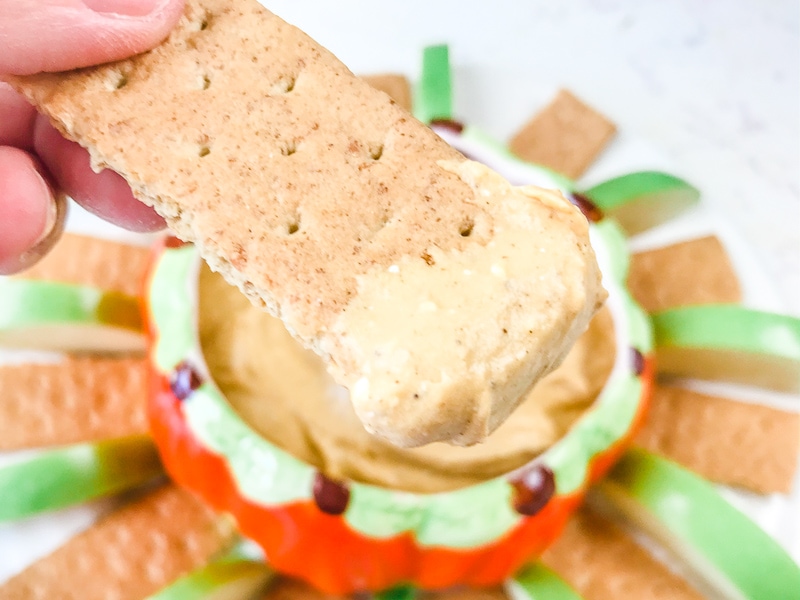 A graham cracker with pumpkin pie dip on the end and a bowl of pumpkin pie cheesecake dip in the background.d