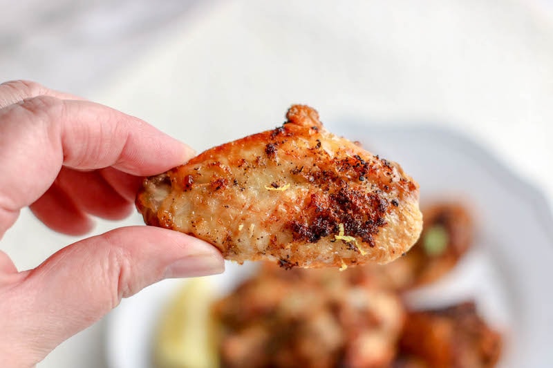 A hand holding a chicken wing above a plate of chicken wings.