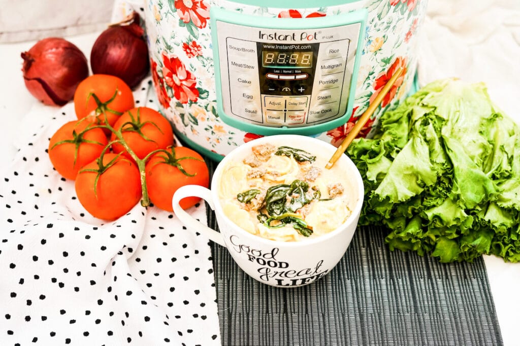 A bowl of tortellini soup in front of an instant pot and next to fresh tomatoes and lettuce leaves.