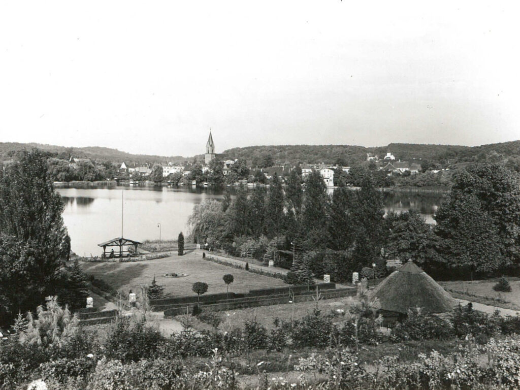 Im Vordergrund sind mehrere Gartenanlagen zu sehen. Von dort aus Blick über den Schermützelsee Richtung Stadt, bei der der große Kirchturm vorne klar zu sehen ist.