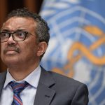 A man with a loosened necktie stands in front of a logo for the World Health Organization.