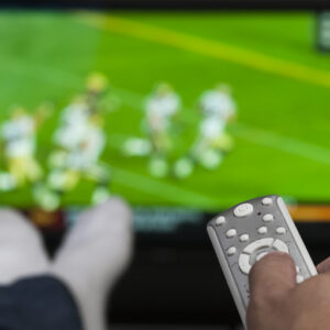 Hand holding a tv remote control. TV has a blurry image of people playing football