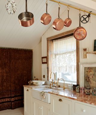 Angled shot of cream lower cabinets in a kitchen with orange stone counters with hanging copper pots on brass rail above and large bright window to the right with lace window dressing