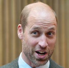 Prince William wearing a white shirt and gray blazer looking surprised standing against a tan wall