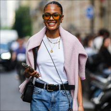  A guest wears orange sunglasses, gold earrings, a white halter-neck tank-top, gold chain pendant necklaces, a pale pink wool pullover, a black shiny leather shoulder bag from Givenchy, a black crossbody camera, a black shiny leather belt, blue faded denim large mom jeans pants, during the Bluemarble show, during Paris Fashion Week Menswear Spring Summer 2023, on June 21, 2022 in Paris, France. (Photo by Edward Berthelot/Getty Images )