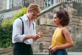 a man (leo woodall as dexter) holds a cigarette and looks down as he stands with a woman (ambika mod as emma) holding a purse