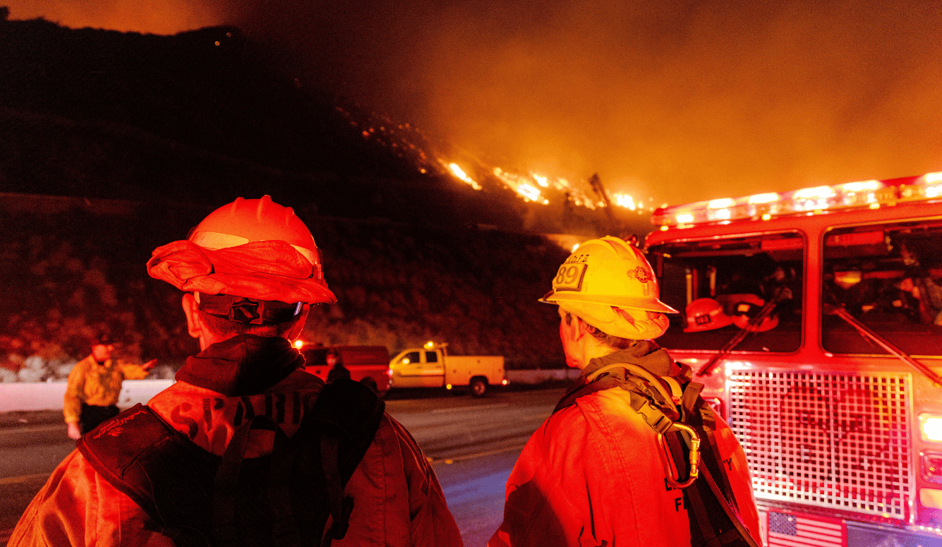 Two firefighters, next to a fire truck, by the side of a highway, across from a blaze. 