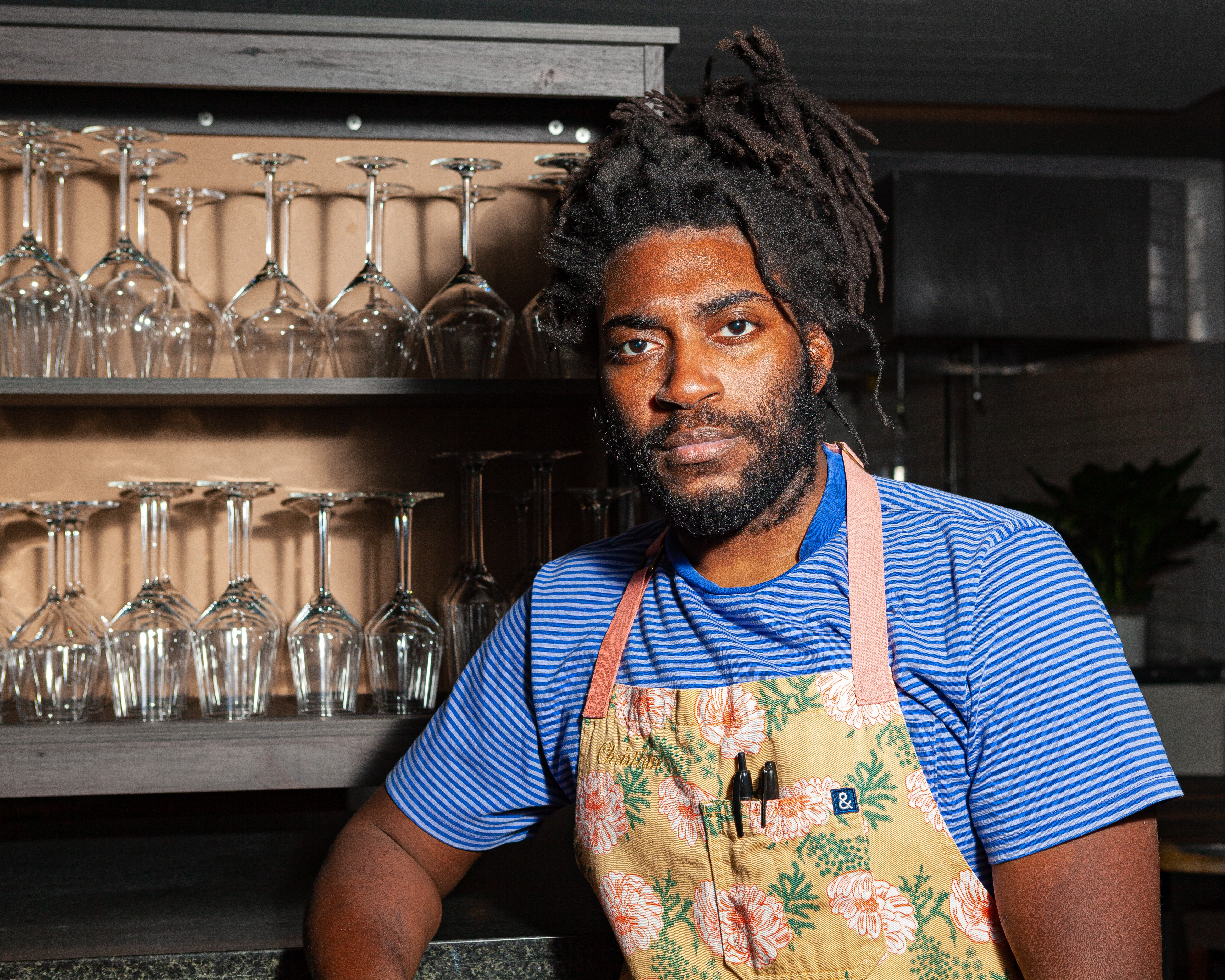 A chef posing near a shelf with glasses.