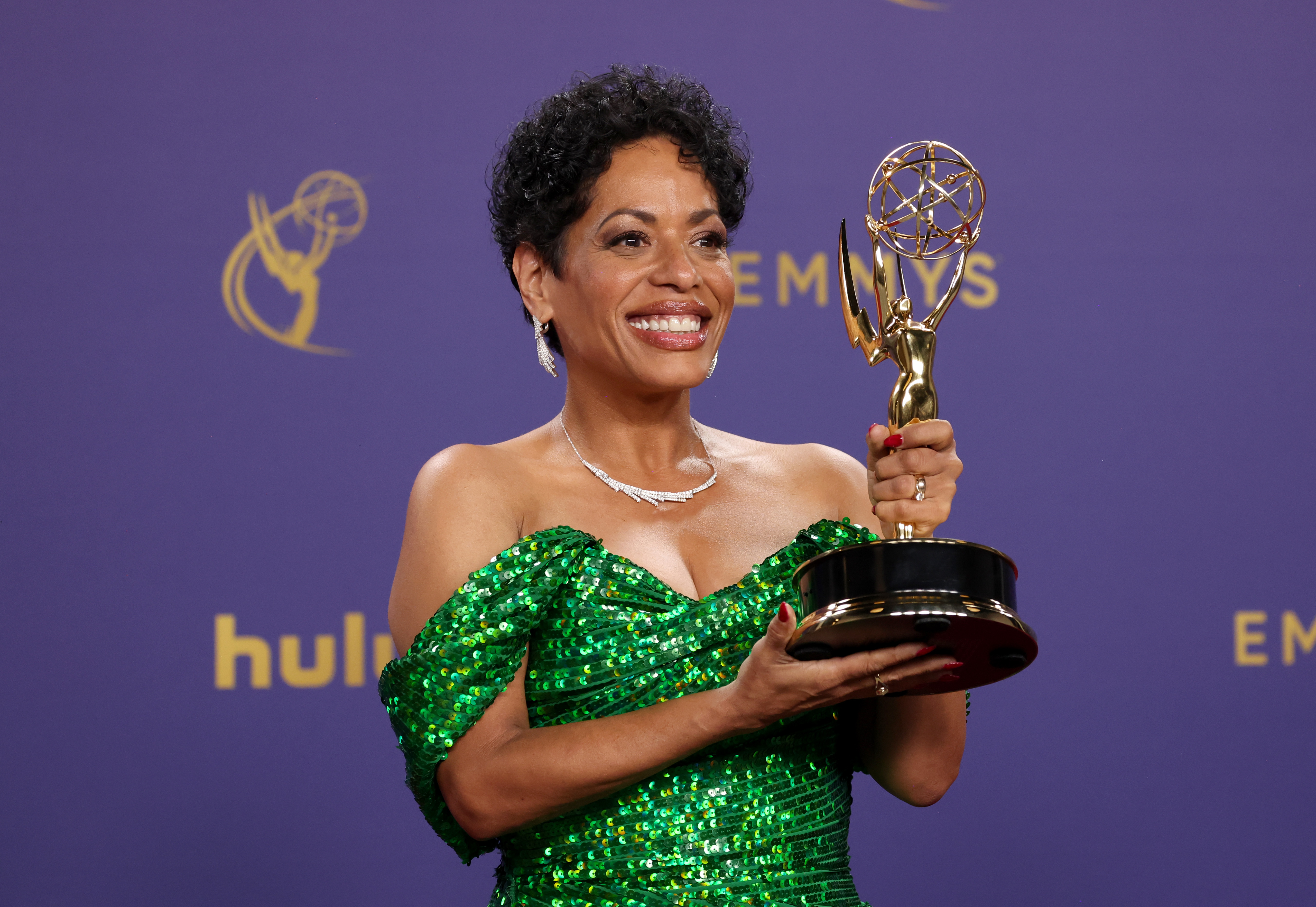 Smiling woman in a sparkling green dress holds up an Emmy Award.