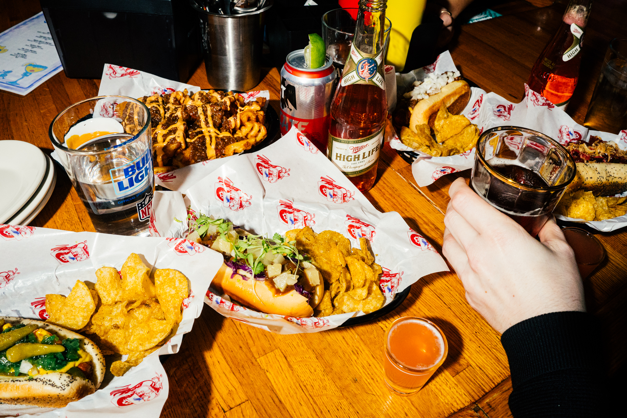 Hot dogs, burgers, beers, and colorful shots crowd a wooden table at a bar.