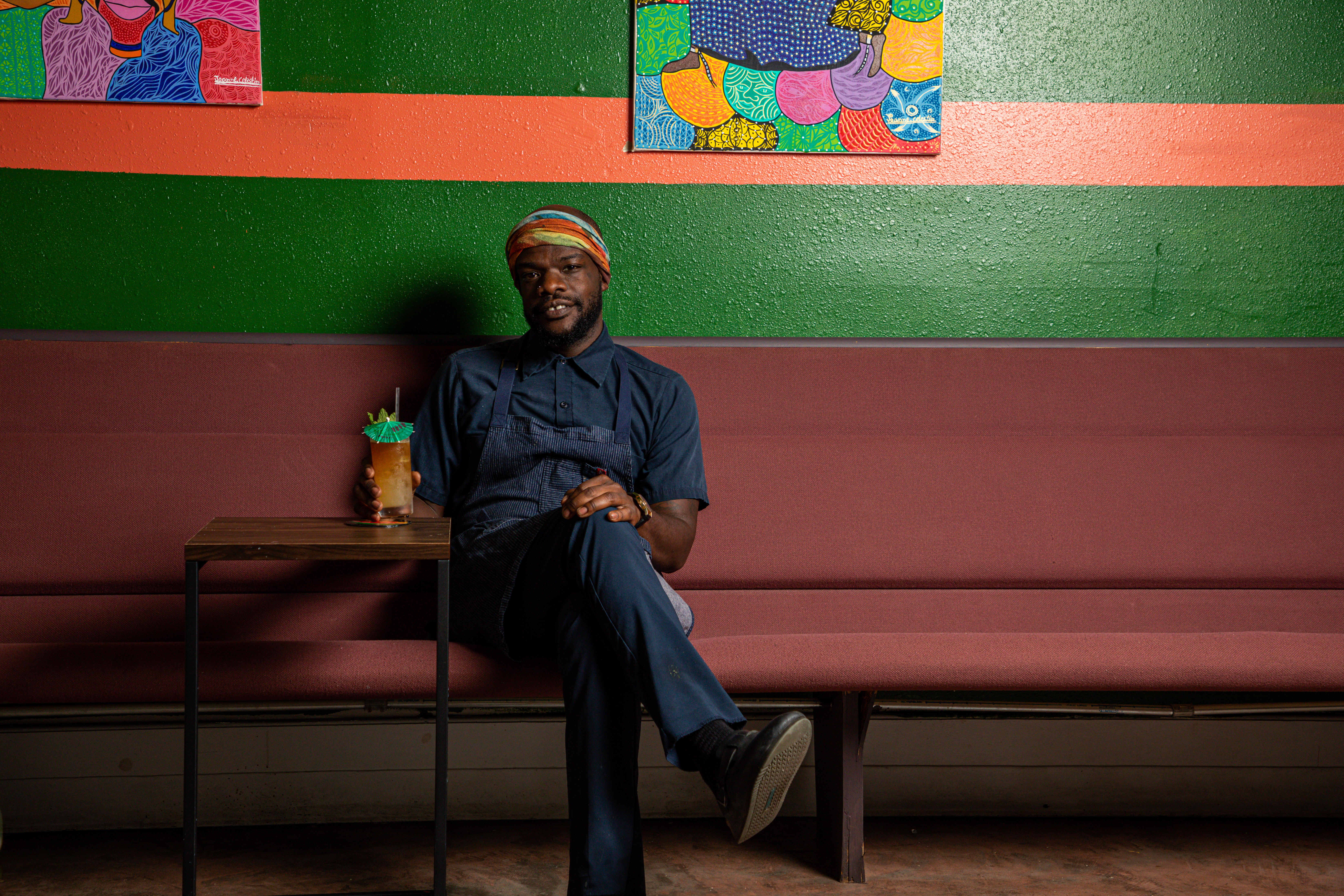 Charly Pierre sits at a mauve banquette in front of a green and pink wall adorned with Haitian art with cocktail in hand, looking at the camera.