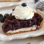 Slice of Blueberry Pie on a white plate topped with whipped cream.