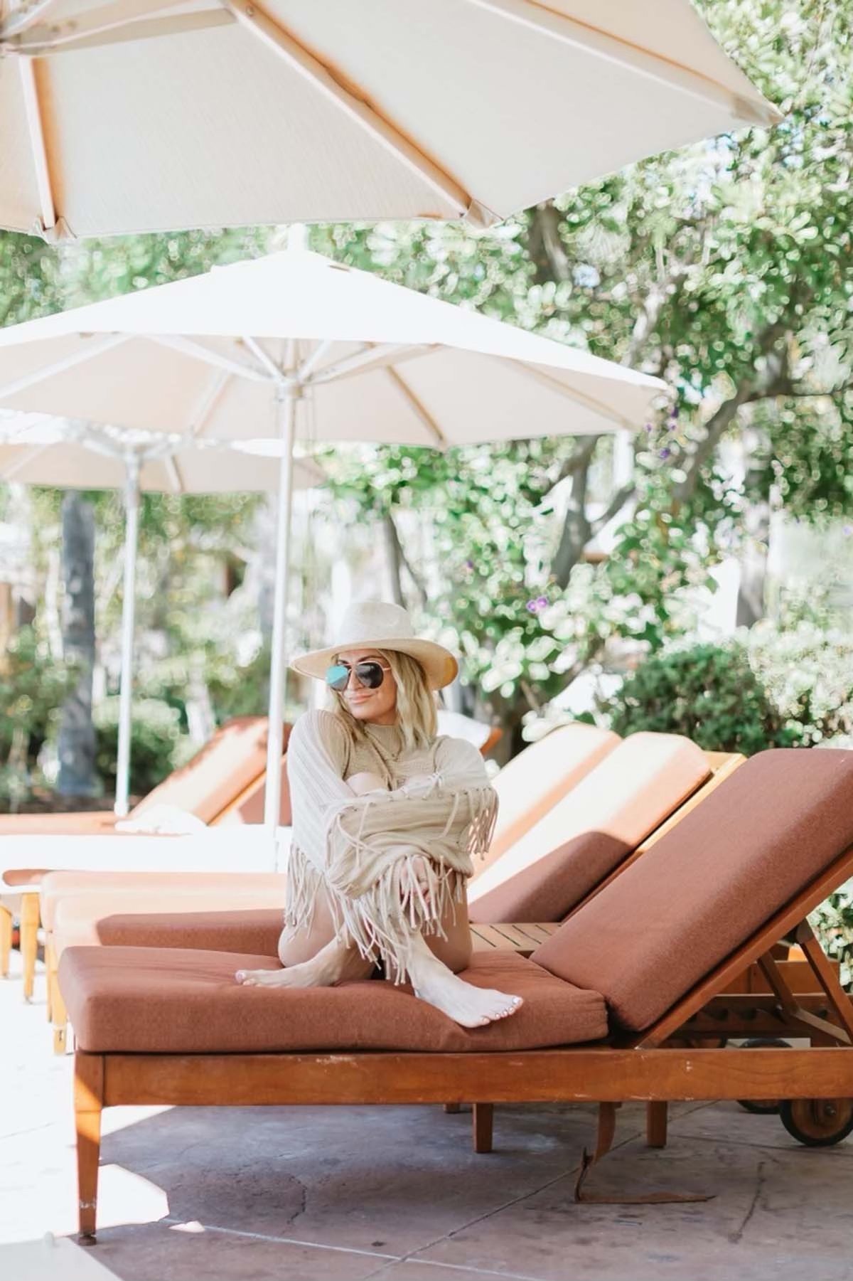 A woman in a hat and sunglasses sits crossed-legged on a lounge chair under a patio umbrella.