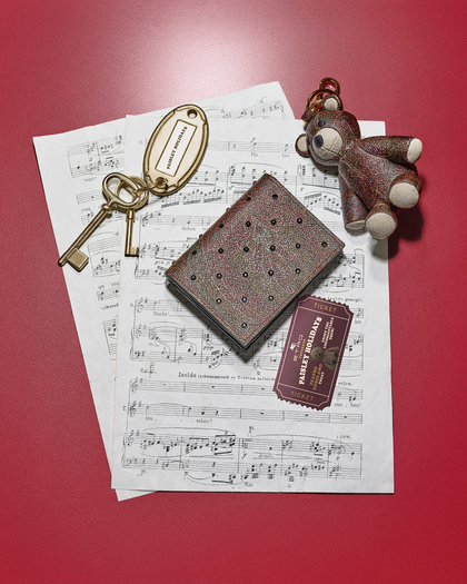 Arrangement of items on a red surface, including a stack of sheet music, a textured Arnica wallet with strass, a vintage-style brass keychain with two keys and an oval tag reading 'Passenger Boarding,' a small teddy bear keychain with a glittery finish, and a red train ticket labeled 'Paisley Holidays.' The scene evokes a sense of travel and nostalgia, with musical notes and keepsakes.