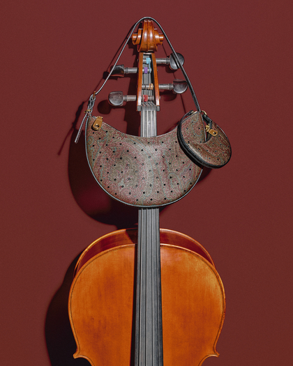 Close-up of a cello standing against a red background, with a ETRO Hobo bag with strass draped over its scroll and pegs. The bag has a curved shape with a Arnica pattern featuring small dots and a matching round coin purse attached. The warm wood tones of the cello contrast with the textured design of the bag, creating a striking visual on the red backdrop.