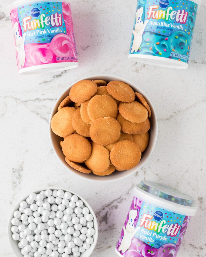 Bowl of vanilla wafers, pink, blue, purple frosting, and bowl of white round candy.