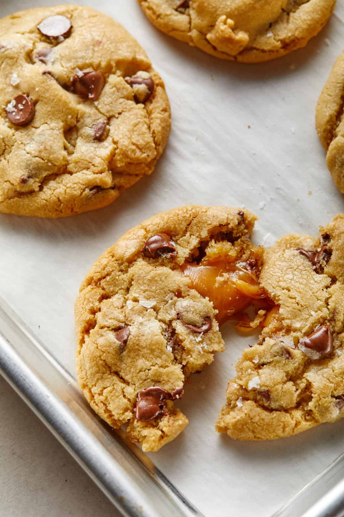 Salted caramel chocolate chip cookies on a baking pan with one broken in half with the caramel oozing out.