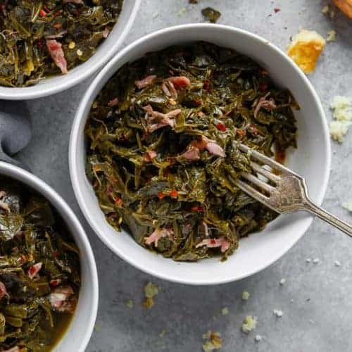 Overhead of three white bowls filled with Southern Collard Greens with hamhock against gray background