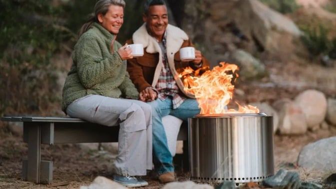 two people sit side by side on a bench behind a solo stove bonfire 2.0. They're both looking at the fire while smiling and both hold white mugs