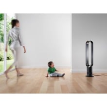 a woman walks across the room to her child who's sitting on the floor in front of a dyson fan