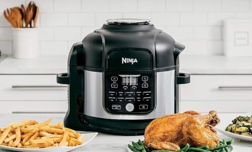 a ninja foodi air fryer and pressure cooker combo sits on a kitchen counter with several dishes of food in front of it including a roast chicken, french fries, and brussel sprouts