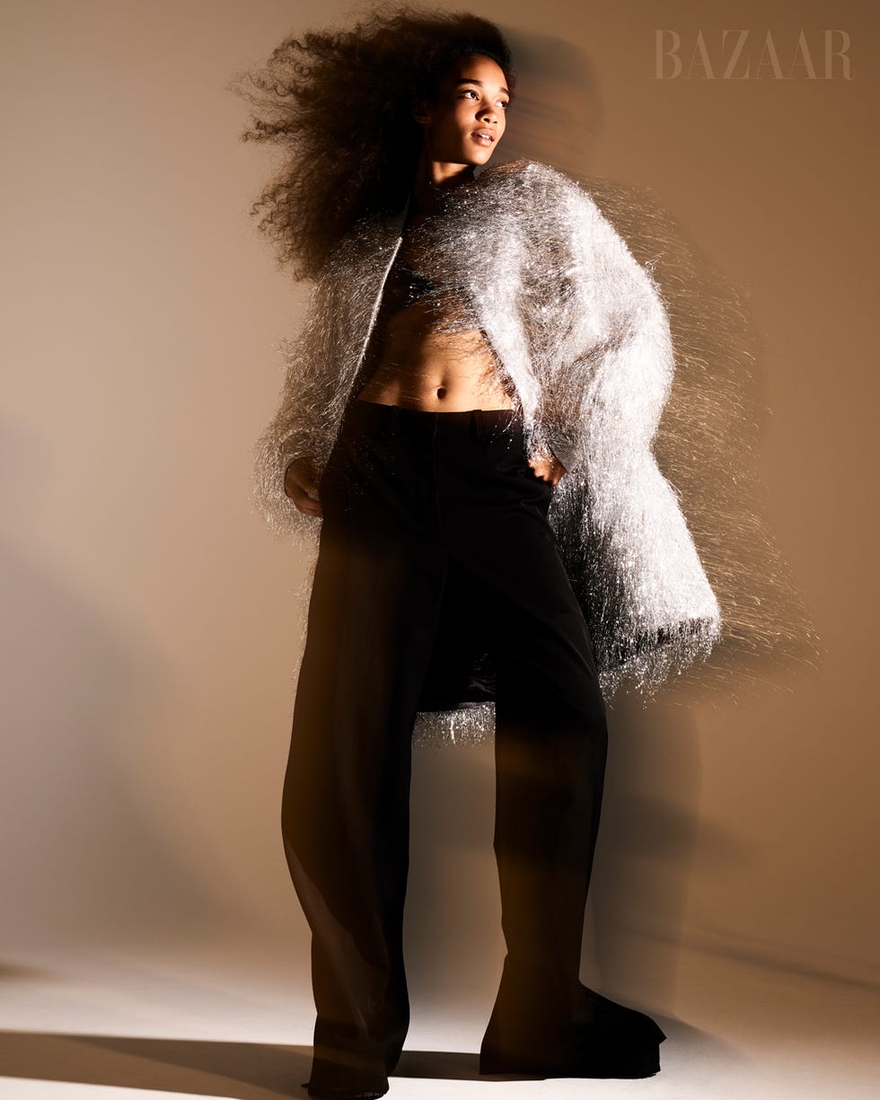 woman standing in white shimmery coat over black crop top and pants in front of tan wall